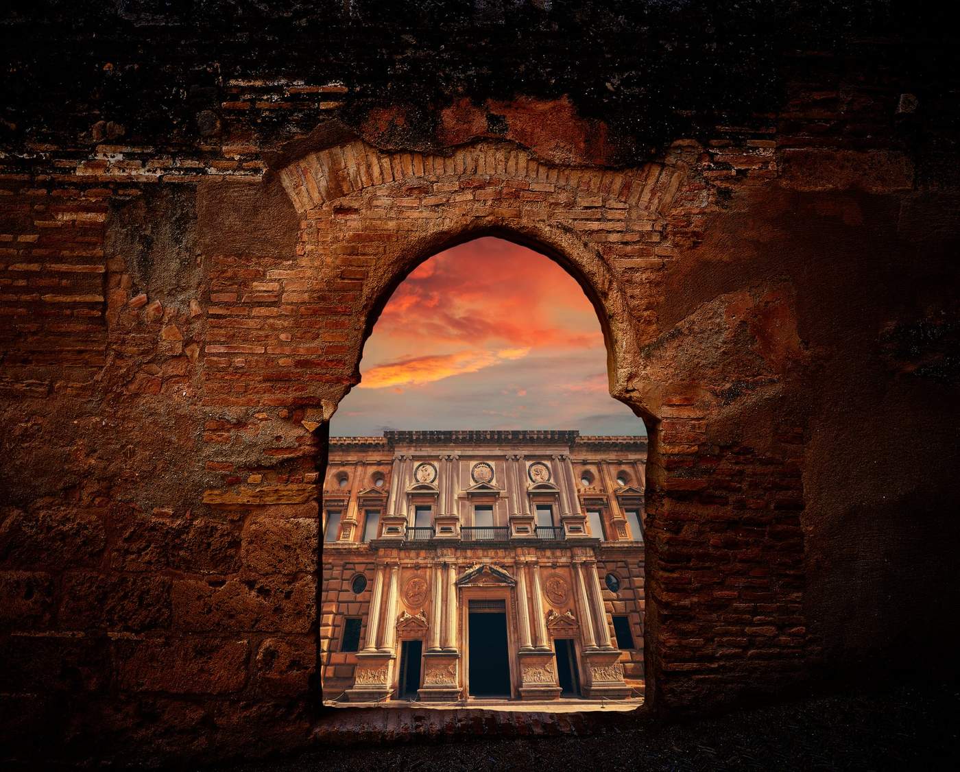 Vue encadrée par une arche en pierre sur un palais de la Renaissance dans l'Alhambra de Grenade, au coucher du soleil, avec un ciel coloré en arrière-plan.