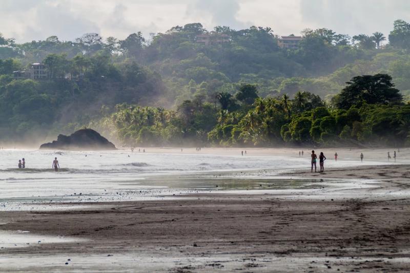 Vue sur la plage de Manuel Antonio avec des palmiers luxuriants en arrière-plan et des personnes profitant de l'eau en bord de mer.