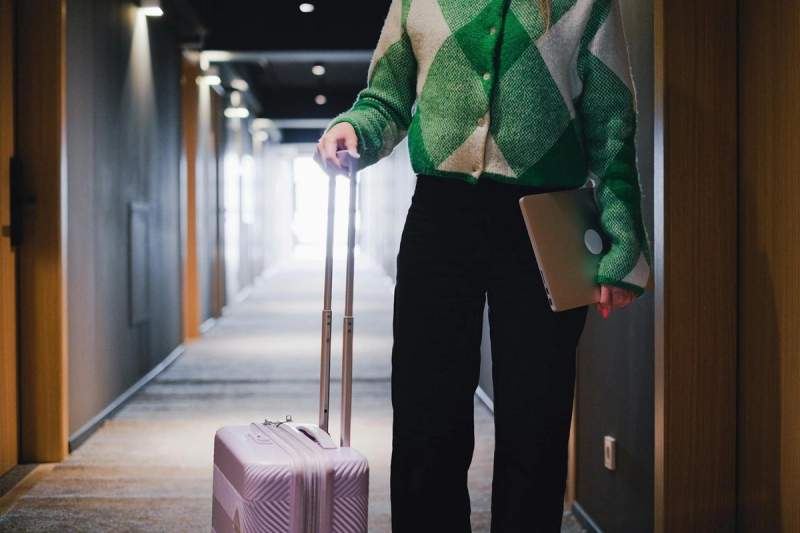 Personne marchant dans un couloir d’hôtel, tenant une valise rose et un ordinateur portable.