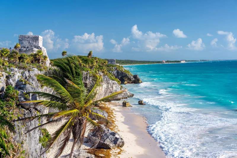 Vue spectaculaire des ruines mayas de Tulum sur une falaise surplombant la mer des Caraïbes, avec des palmiers, des plages de sable fin, et des eaux turquoise étincelantes sous un ciel bleu dégagé.