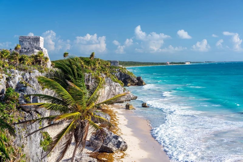 Vue spectaculaire des ruines mayas de Tulum sur une falaise surplombant la mer des Caraïbes, avec des palmiers, des plages de sable fin, et des eaux turquoise étincelantes sous un ciel bleu dégagé.