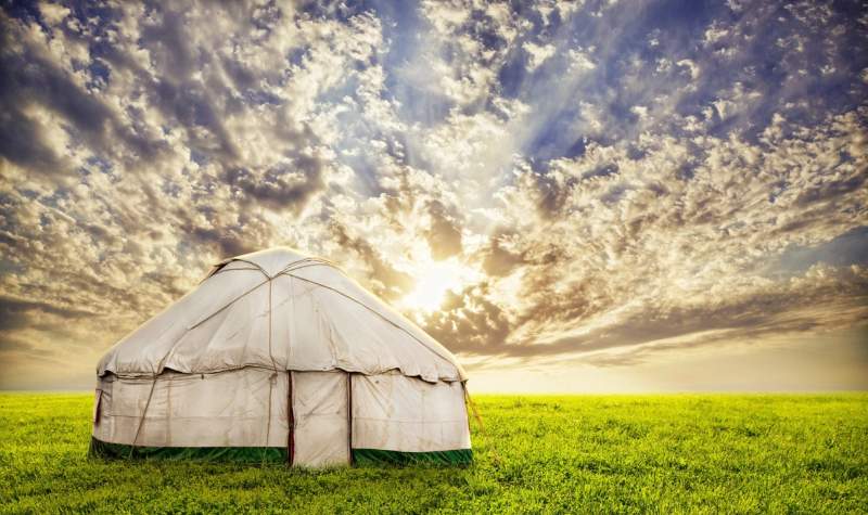 Yourte traditionnelle en toile blanche installée au milieu d’un champ verdoyant, avec un ciel nuageux et un soleil couchant à l’horizon.