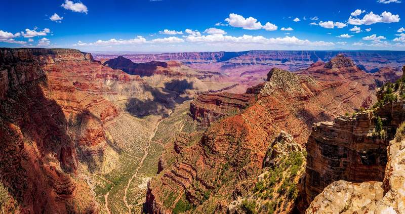 Vue panoramique du Grand Canyon avec ses falaises rocheuses striées de rouge et d'ocre sous un ciel bleu parsemé de nuages