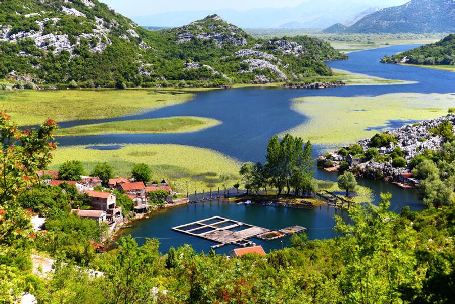 Vue plongeante sur un petit village et ses bassins de pisciculture au bord du lac Skadar, entouré de nénuphars et de collines calcaires au Monténégro