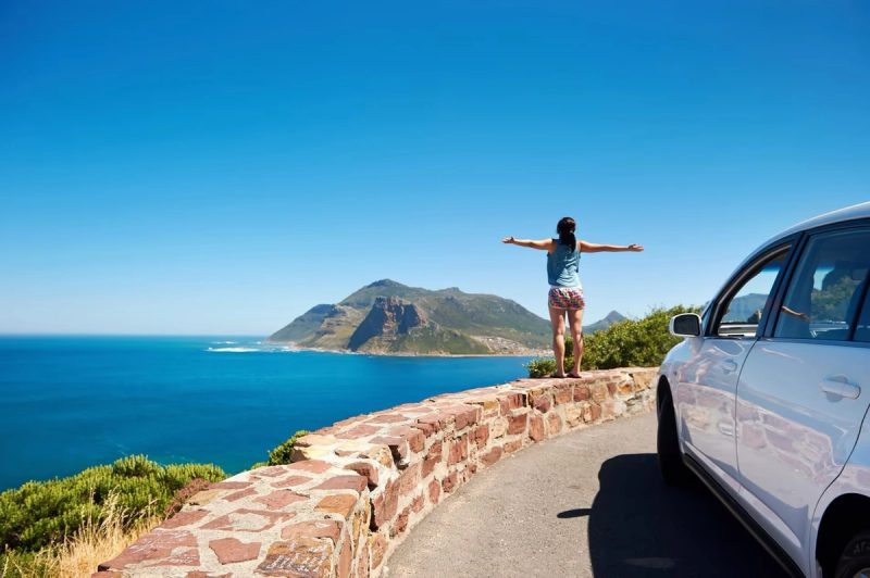 Personne debout les bras écartés sur un muret surplombant une route côtière, face à un paysage de mer turquoise et de montagnes sous un ciel bleu clair, avec une voiture blanche garée à droite.