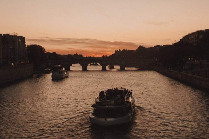 Bateaux naviguant sur la Seine à Paris au coucher du soleil, avec le Pont Neuf en arrière-plan et un ciel aux tons orangés