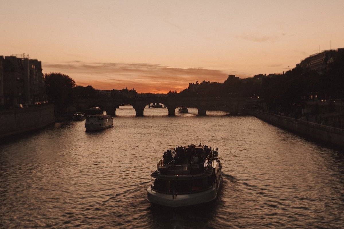 Bateaux naviguant sur la Seine à Paris au coucher du soleil, avec le Pont Neuf en arrière-plan et un ciel aux tons orangés