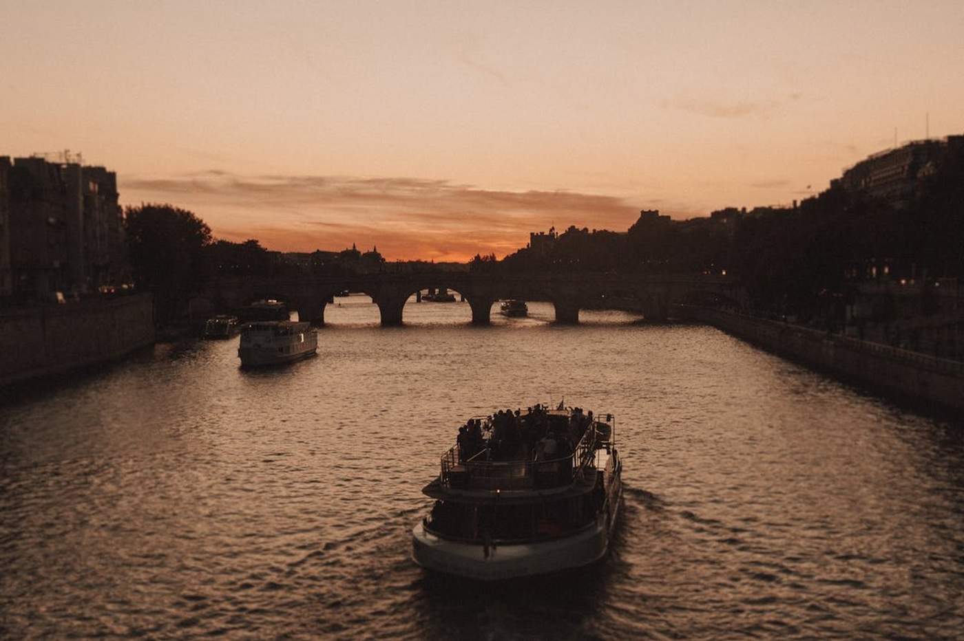 Bateaux naviguant sur la Seine à Paris au coucher du soleil, avec le Pont Neuf en arrière-plan et un ciel aux tons orangés
