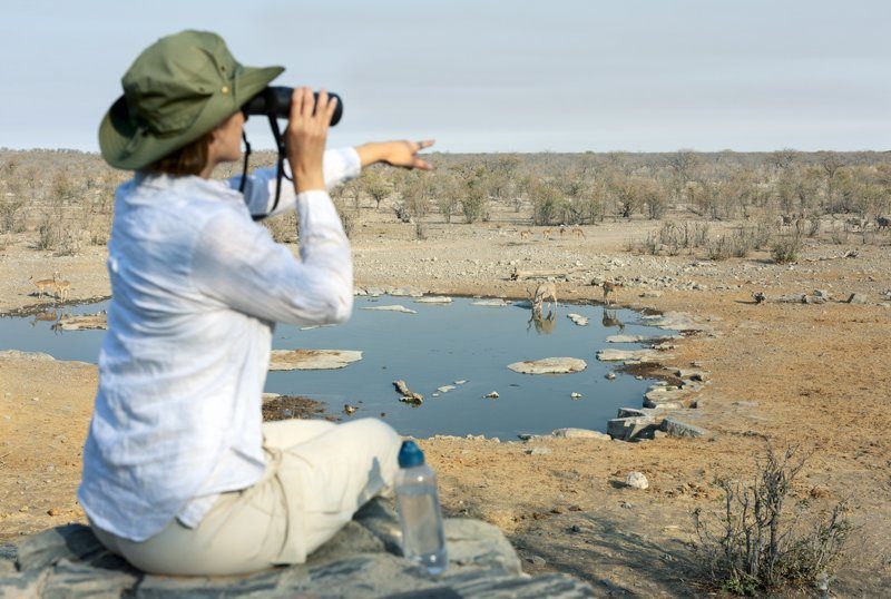 Une femme portant un chapeau de safari observe des animaux à l’aide de jumelles près d’un point d’eau dans une savane aride