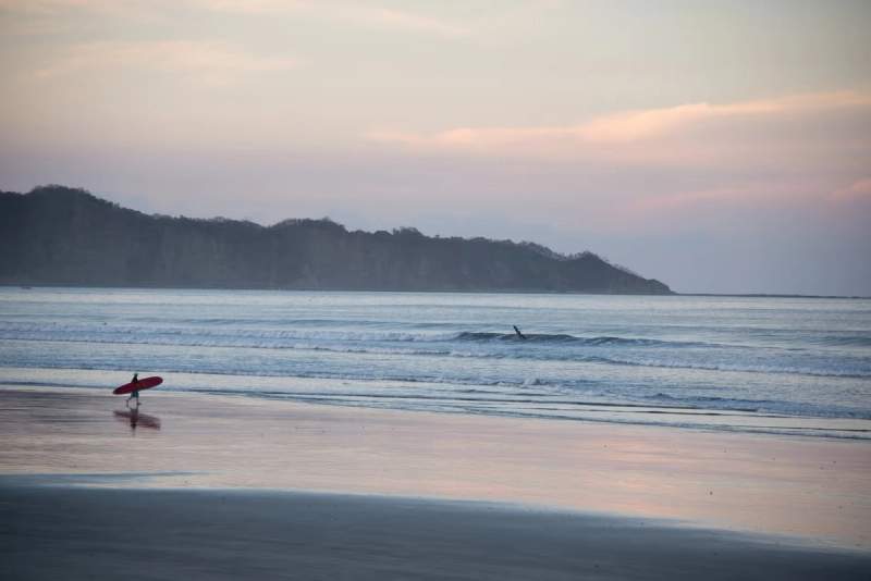 Un surfeur marchant avec sa planche le long d'une plage calme, avec des vagues douces et des collines en arrière-plan, au coucher du soleil.