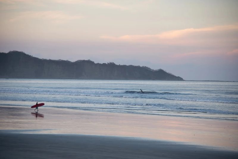Un surfeur marchant avec sa planche le long d'une plage calme, avec des vagues douces et des collines en arrière-plan, au coucher du soleil.