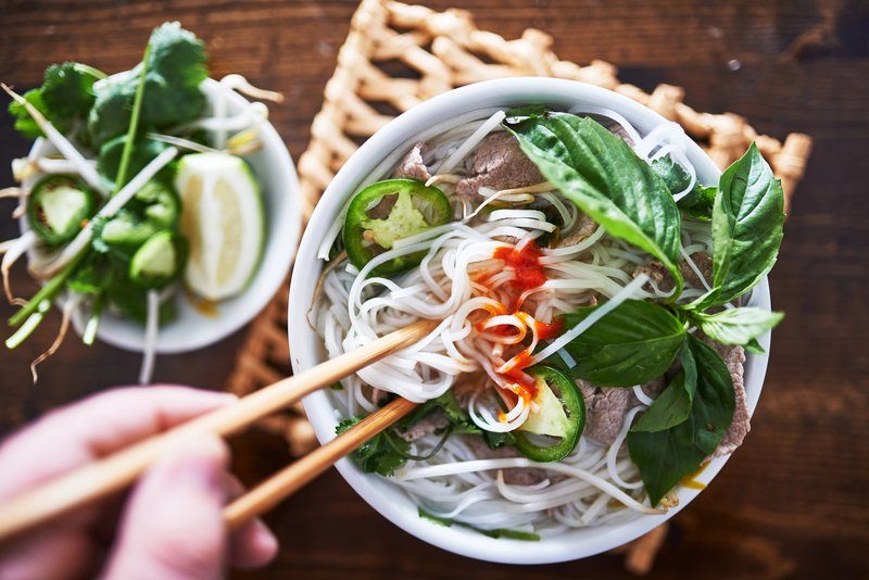 Bol de phở traditionnel vietnamien garni de bœuf, d’herbes fraîches et de piment, avec baguettes en main.