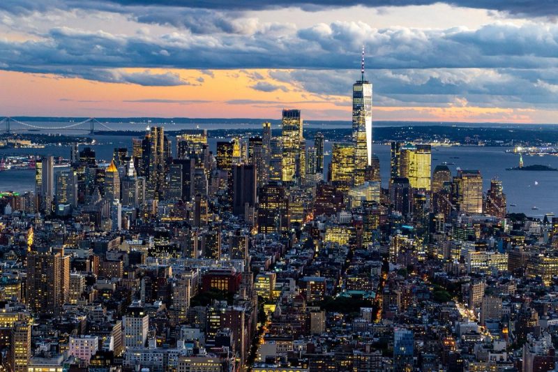 Vue panoramique de la ville de New York au crépuscule, avec des gratte-ciels illuminés et un ciel nuageux.