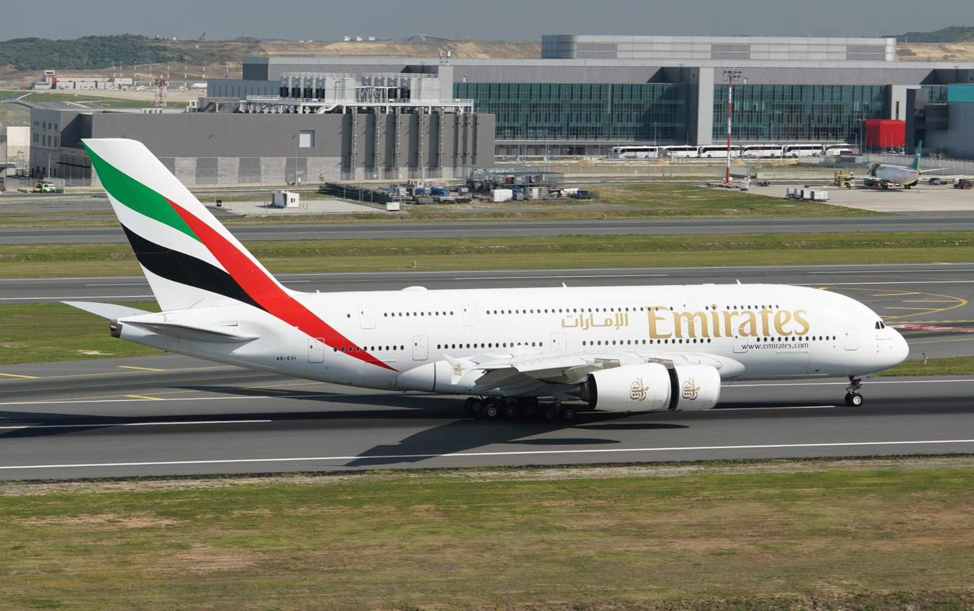 Avion Airbus A380 d'Emirates sur la piste d'un aéroport, prêt pour le décollage, avec un arrière-plan de bâtiments modernes de l'aéroport.