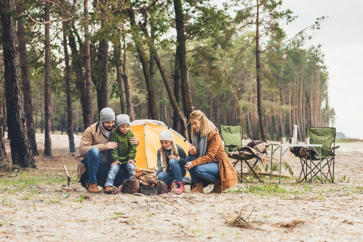 Famille en plein air installant un campement, avec deux enfants et leurs parents assis autour d’un petit feu devant une tente dans une forêt.