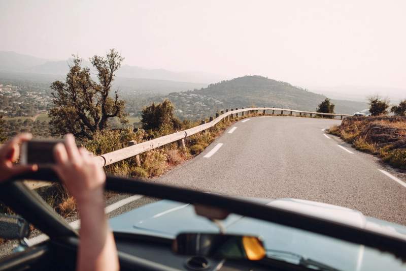 Vue depuis une voiture décapotable sur une route de campagne sinueuse en France, avec collines et végétation typiquement méditerranéenne à l’arrière-plan.