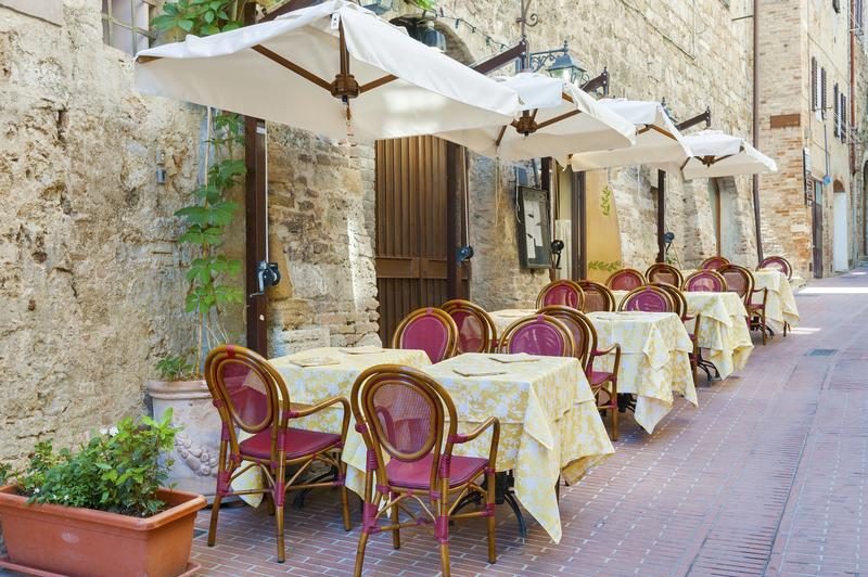 Terrasse d’un restaurant typique à San Gimignano, avec des chaises en rotin rouge et des nappes jaunes, le long d’une ruelle en pierre.