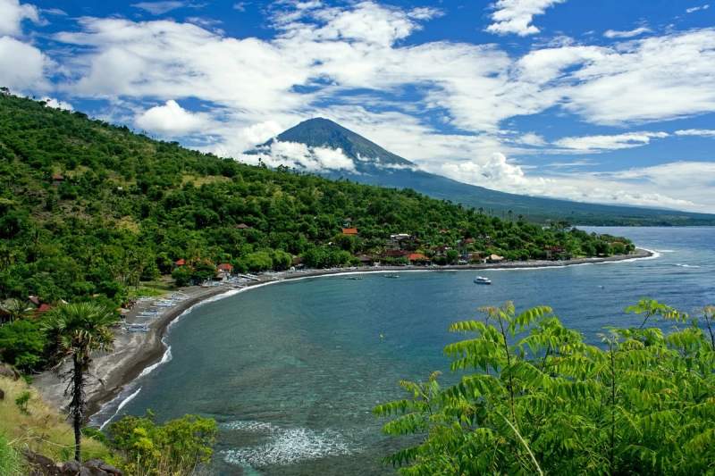 Vue panoramique de la baie d'Amed, Bali, avec la plage, des bateaux traditionnels et le Mont Agung en arrière-plan, sous un ciel bleu partiellement nuageux.