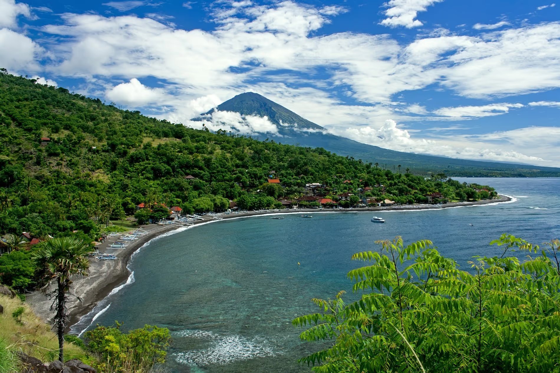 Vue panoramique de la baie d'Amed, Bali, avec la plage, des bateaux traditionnels et le Mont Agung en arrière-plan, sous un ciel bleu partiellement nuageux.