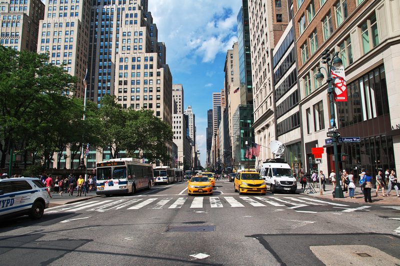 Vue d’une large avenue à New York, avec des taxis jaunes emblématiques au centre, des bus, des bâtiments de grande hauteur, et des piétons marchant le long des trottoirs par une journée ensoleillée.