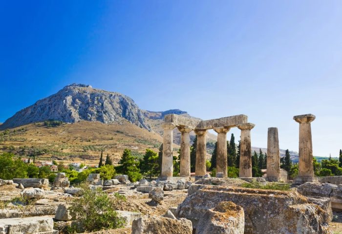 Ruines du temple d'Apollon à Corinthe avec une montagne en arrière-plan sous un ciel bleu.