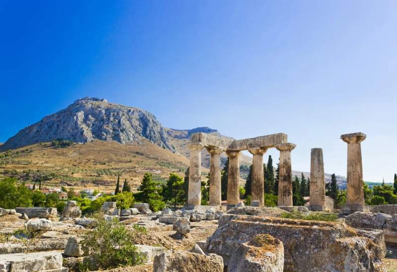 Ruines du temple d'Apollon à Corinthe avec une montagne en arrière-plan sous un ciel bleu.