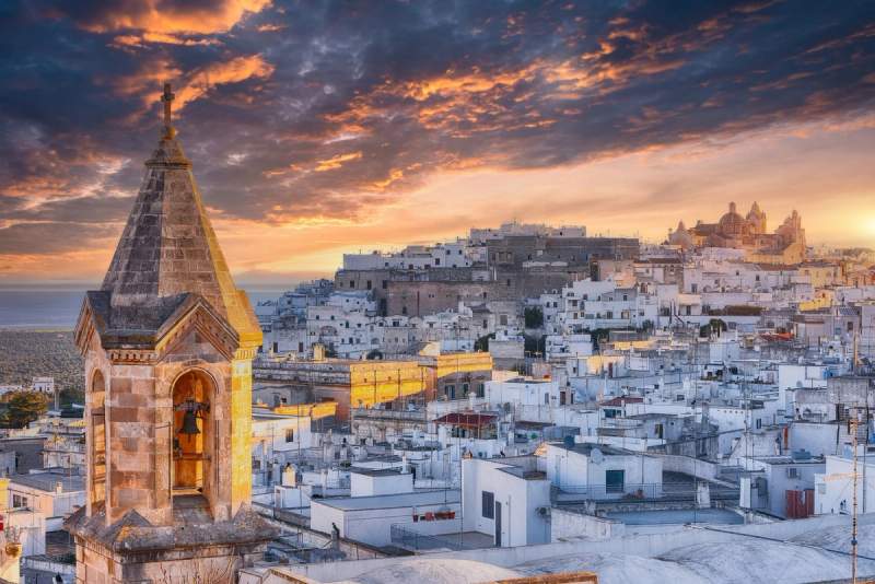 Vue panoramique d'Ostuni au coucher du soleil, avec ses maisons blanches et une cloche en premier plan. 