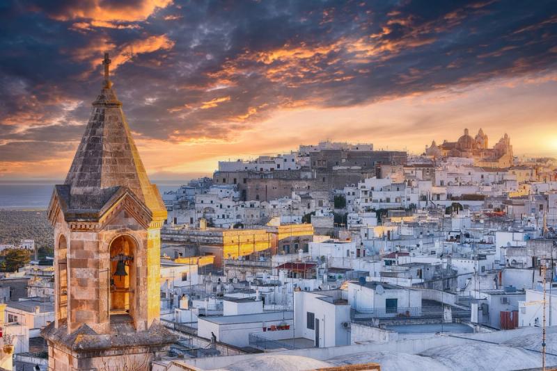 Vue panoramique d'Ostuni au coucher du soleil, avec ses maisons blanches et une cloche en premier plan.