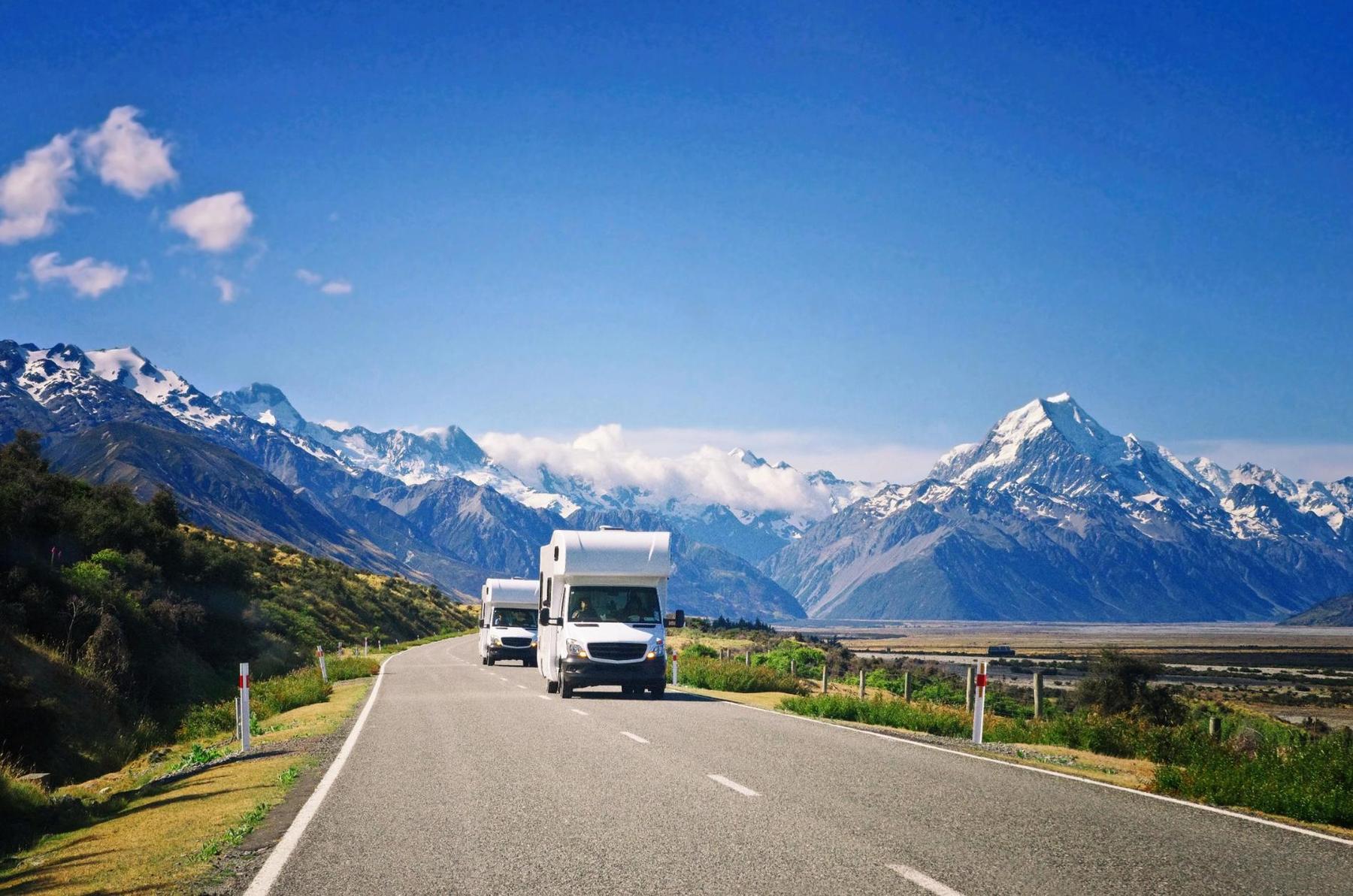 Deux camping-cars parcourant une route de montagne en Nouvelle-Zélande, entourés de paysages spectaculaires avec des sommets enneigés sous un ciel bleu clair.