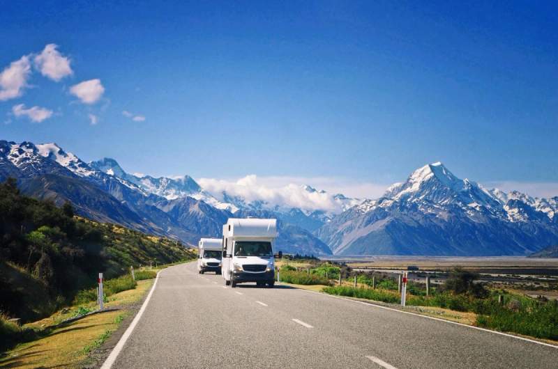 Deux camping-cars parcourant une route de montagne en Nouvelle-Zélande, entourés de paysages spectaculaires avec des sommets enneigés sous un ciel bleu clair.