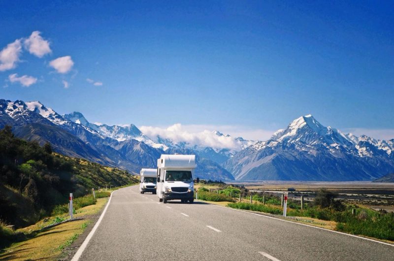 Deux camping-cars parcourant une route de montagne en Nouvelle-Zélande, entourés de paysages spectaculaires avec des sommets enneigés sous un ciel bleu clair.