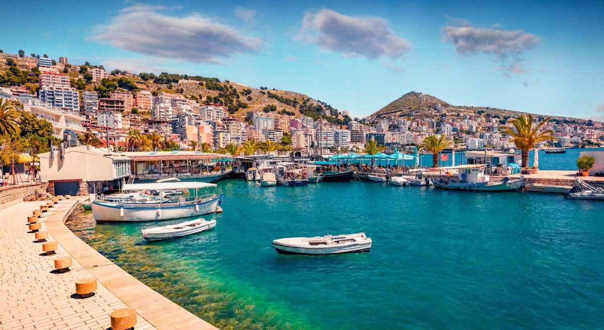 Vue sur le port de Saranda, en Albanie, avec ses bateaux amarrés et la ville en arrière-plan, entourée de collines et de la mer Ionienne.