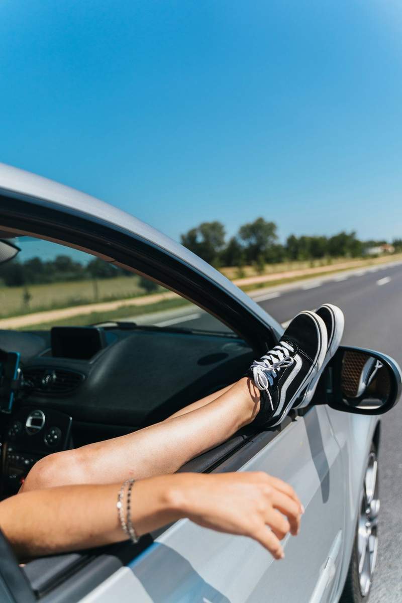 Personne profitant d’un road trip, jambes sorties par la fenêtre d’une voiture sur une route ensoleillée