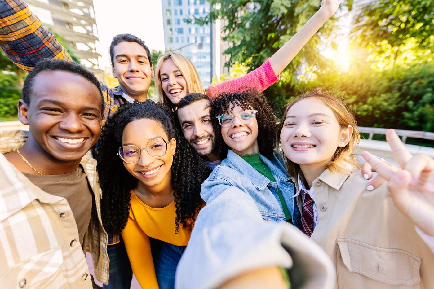Groupe d'amis souriants prenant un selfie en extérieur avec des bâtiments et de la verdure en arrière-plan.