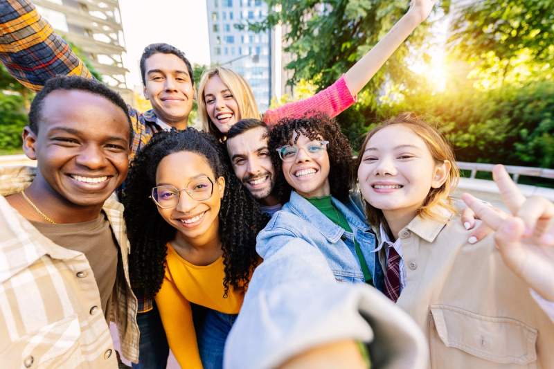 Groupe d'amis souriants prenant un selfie en extérieur avec des bâtiments et de la verdure en arrière-plan.