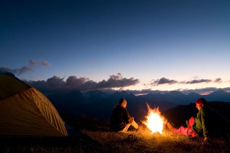 Deux campeurs assis autour d’un feu au coucher du soleil, avec une tente et des montagnes en arrière-plan.