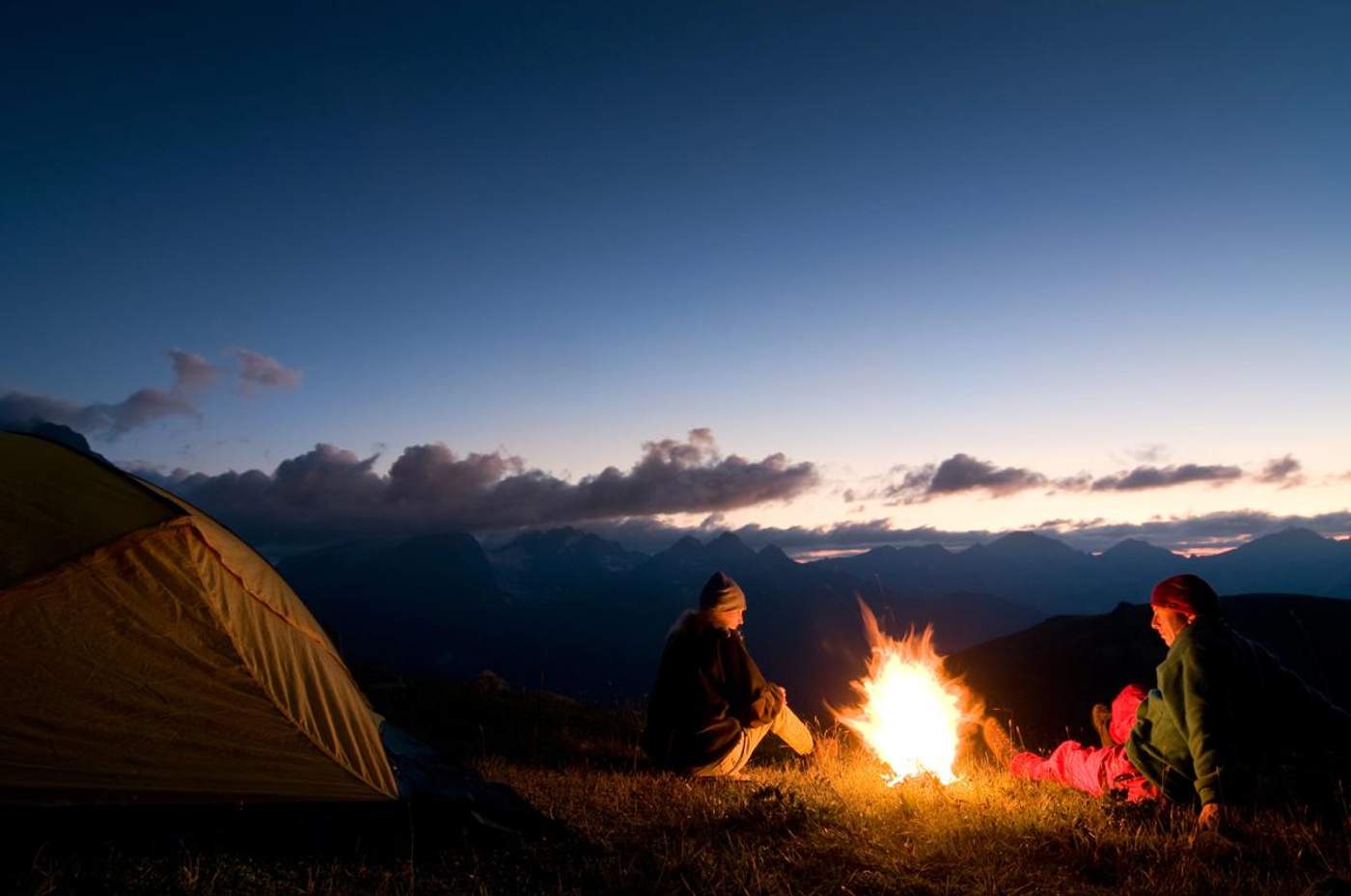 Deux campeurs assis autour d’un feu au coucher du soleil, avec une tente et des montagnes en arrière-plan.