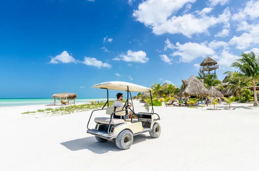 Une voiturette de golf blanche se déplace sur une plage de sable blanc à Holbox, sous un ciel bleu clair, avec des palmiers et des cabanes en arrière-plan.