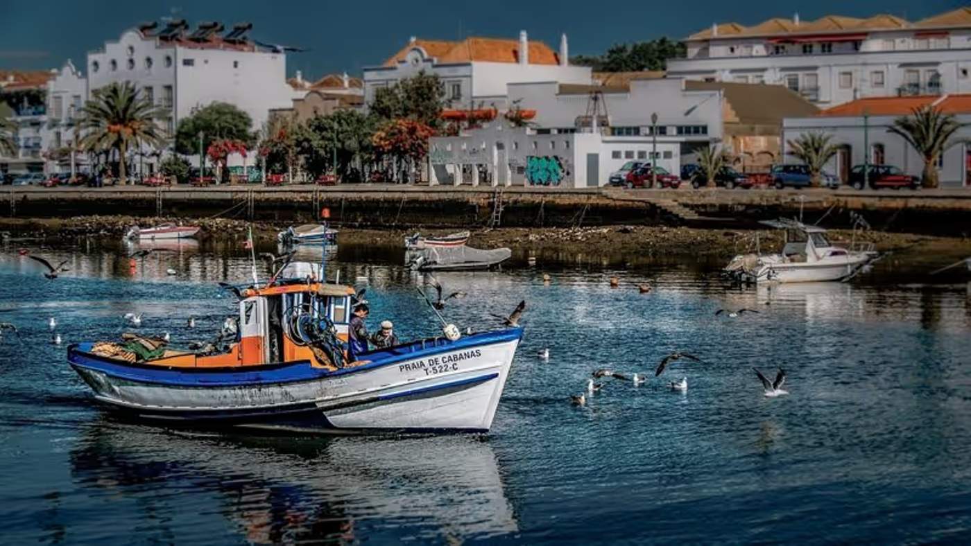 Bateau de pêche naviguant dans le port de Tavira avec des bâtiments en arrière-plan, Portugal.