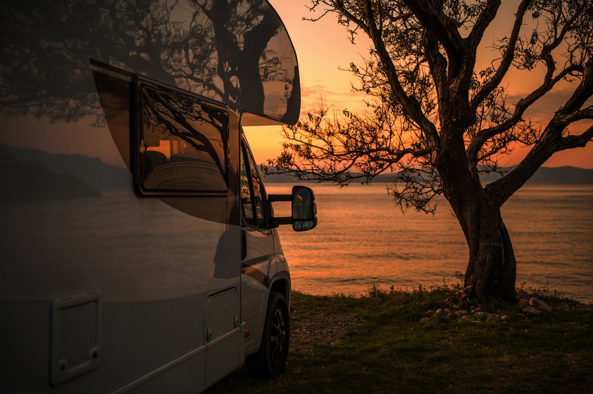 Camping-car stationné face à la mer au coucher du soleil, reflet doré sur la carrosserie et arbre en premier plan.