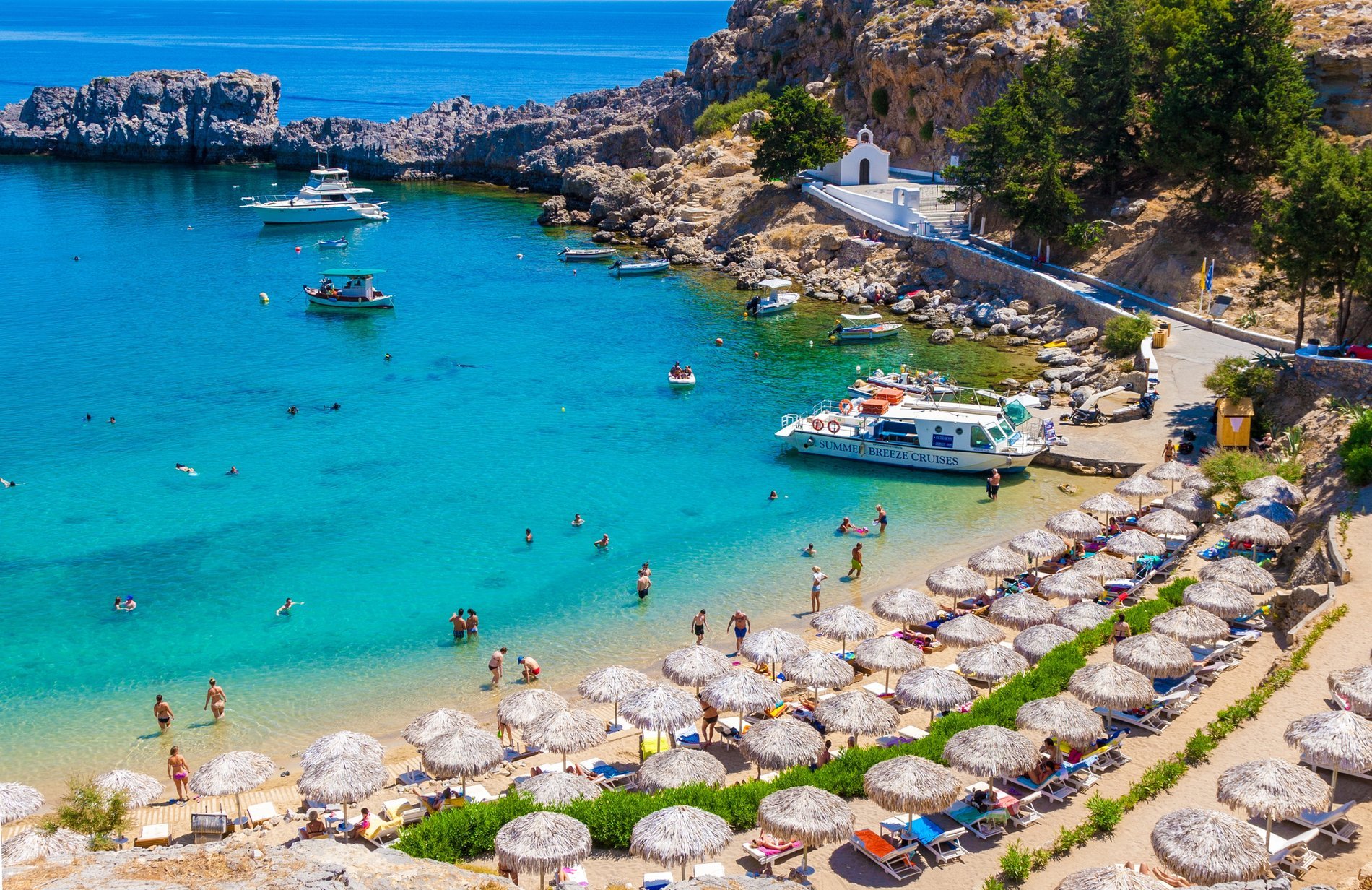 Plage paradisiaque à Rhodes avec parasols en paille, transats et eau turquoise, des baigneurs profitant du soleil et des bateaux ancrés dans une crique rocheuse.