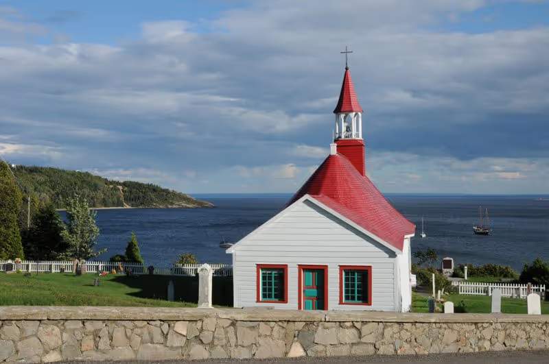 La chapelle historique de Tadoussac avec son toit rouge distinctif, surplombant le fleuve Saint-Laurent et ses voiliers.