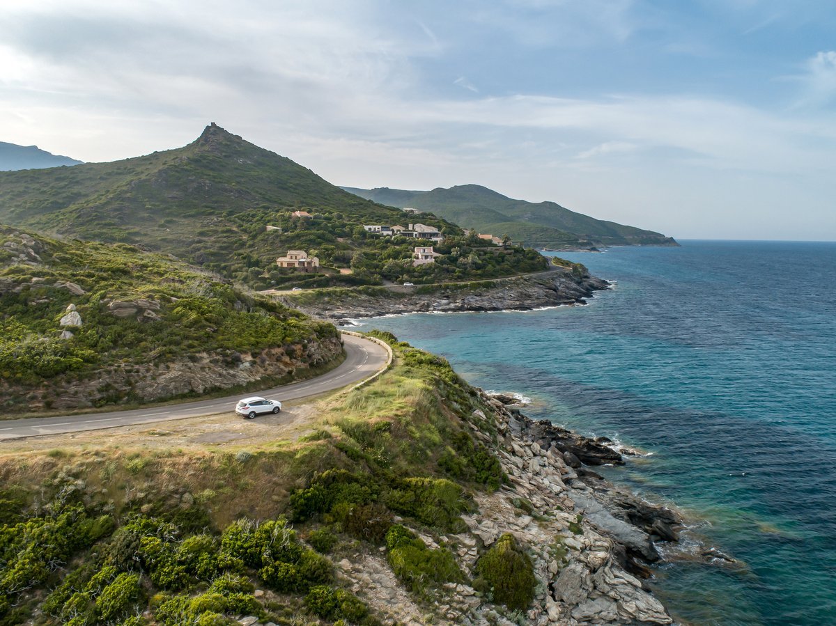 Vue aérienne d'une route côtière sinueuse en Corse longeant la mer Méditerranée, avec une voiture blanche, des collines verdoyantes et des maisons typiques nichées dans le maquis.