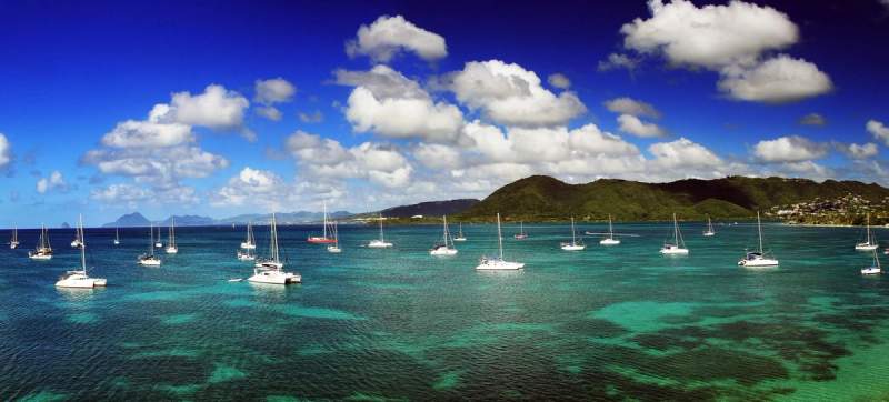 Vue panoramique de la Martinique avec des voiliers sur une mer turquoise sous un ciel bleu et des collines en arrière-plan.