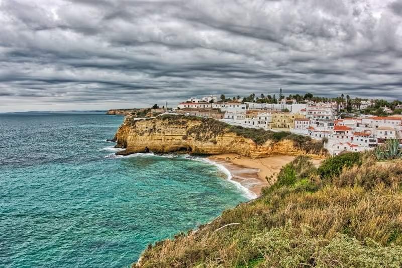 Vue de la plage de Carvoeiro avec ses falaises dorées et ses maisons blanches sous un ciel nuageux.