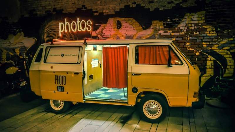 Van photobus jaune transformé en cabine photo mobile, stationné devant un mur de graffitis avec rideaux rouges.