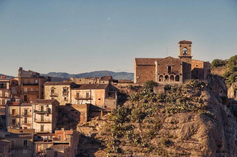Vue pittoresque d'un village sicilien au coucher du soleil, mettant en valeur l'architecture ancienne et les paysages rocheux environnants. 