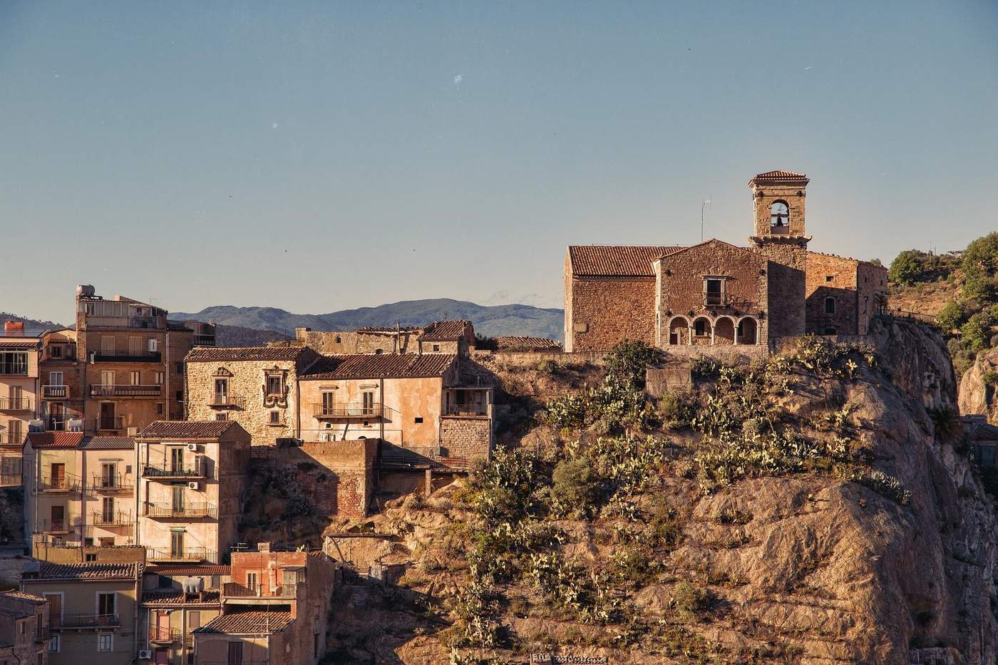 Vue pittoresque d'un village sicilien au coucher du soleil, mettant en valeur l'architecture ancienne et les paysages rocheux environnants.