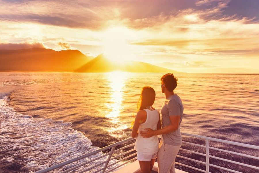 Couple romantique observant un coucher de soleil doré depuis le pont d’un bateau de croisière, avec l’horizon et les montagnes au loin.