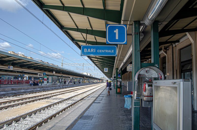 Quai de la gare de Bari Centrale dans les Pouilles, avec les voies ferrées vides et le panneau du quai numéro 1 sous un ciel bleu.